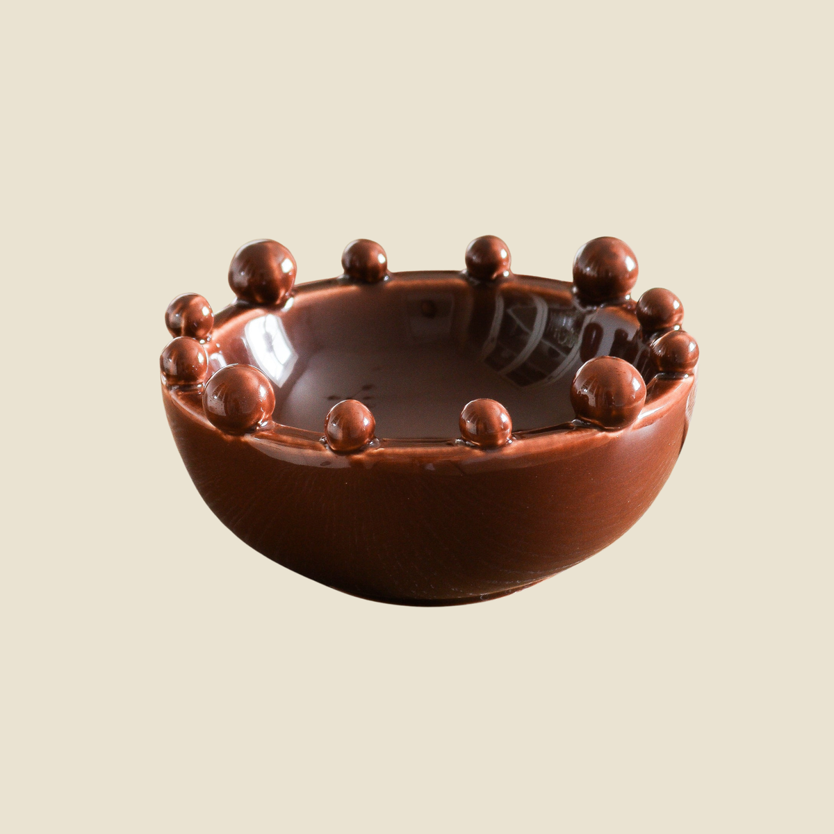 A small glossy brown ceramic bowl with raised spherical details around the rim, shown on a plain warm beige background.