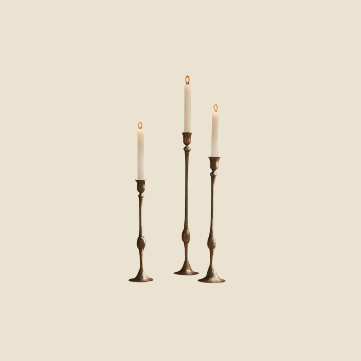 Three vintage-style brass candlestick holders with white taper candles, arranged against a solid light beige background.