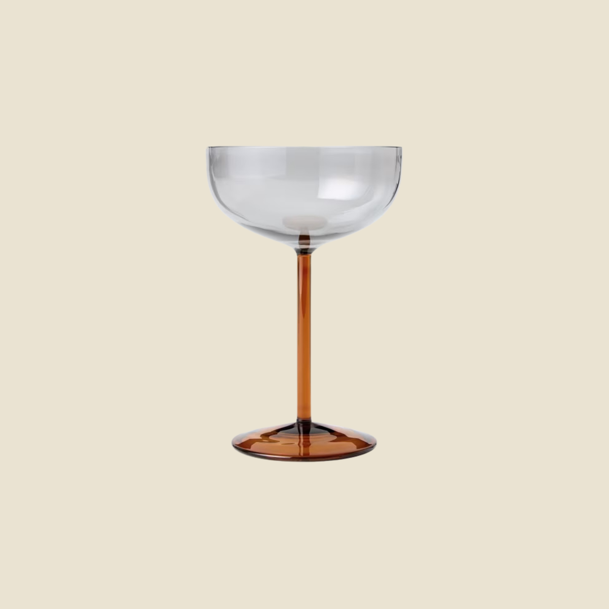 A clear glass coupe glass with a translucent amber-brown stem and base, centered on a solid light beige background.
