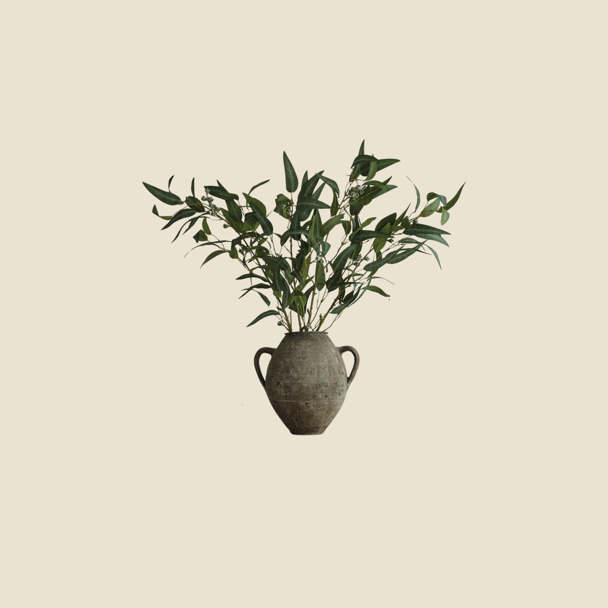 An artificial eucalyptus branch with green leaves and small buds, arranged in a rustic, textured gray ceramic vase with two side handles, set against a solid beige background.