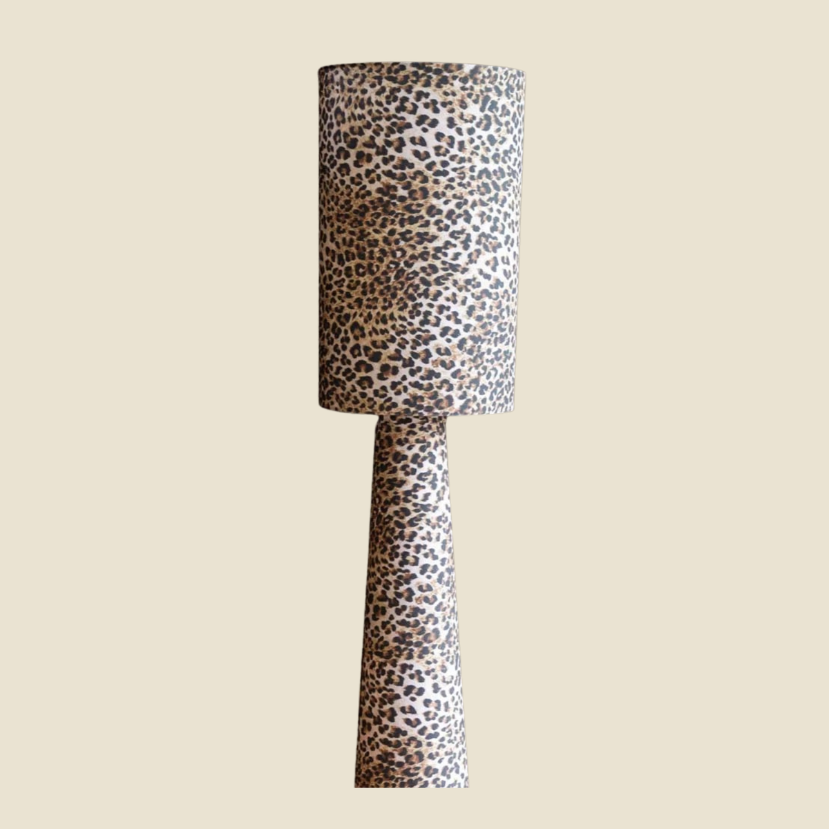 A table lamp with a tapered drum shade and matching tapered base, both covered in a dense leopard print pattern of black and dark brown spots on a warm beige background, set against a solid light beige backdrop.