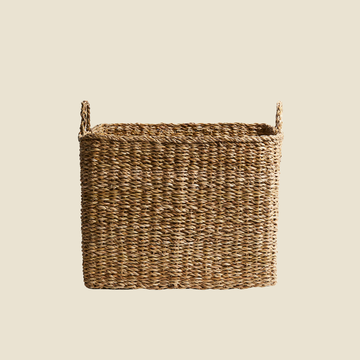 A rectangular woven storage basket made from natural brown seagrass or wicker, featuring two loop handles on the sides, shown against a solid light beige background.